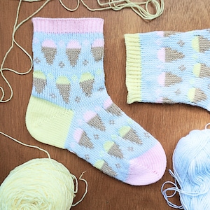 May include: A light blue knitted sock with a pattern of ice cream cones in brown, pink, and green. The heel and toe are pink and yellow, with a pink cuff. Yarn in various colors is scattered around the sock.