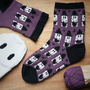 May include: A pair of knitted socks featuring a purple base with a repeating pattern of white and black ghost figures, complemented by black cuffs, heels, and toes. A white fuzzy fabric piece and a ball of purple yarn are positioned nearby.
