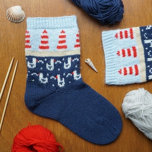 Op de afbeelding: Een handgebreide blauwe sok met een rood-wit vuurtorenpatroon. De sok heeft een marineblauwe voet en een lichtblauwe boord met rood-witte vuurtorenontwerpen. Extra gebreide stukken en garen zijn zichtbaar.
