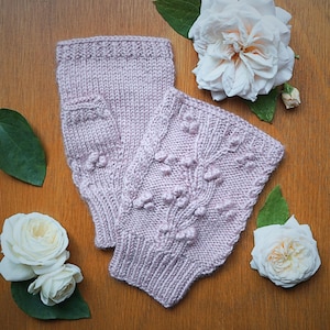May include: A pair of light purple knitted fingerless gloves with a textured floral design. The gloves are made of soft yarn and are displayed on a wooden surface, with white roses and green leaves.