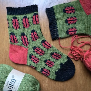 May include: Hand-knitted green socks featuring a repeating pattern of red and black ladybirds. The heel and toe are red, and the cuff is black. A ball of green and red yarn are in the background.
