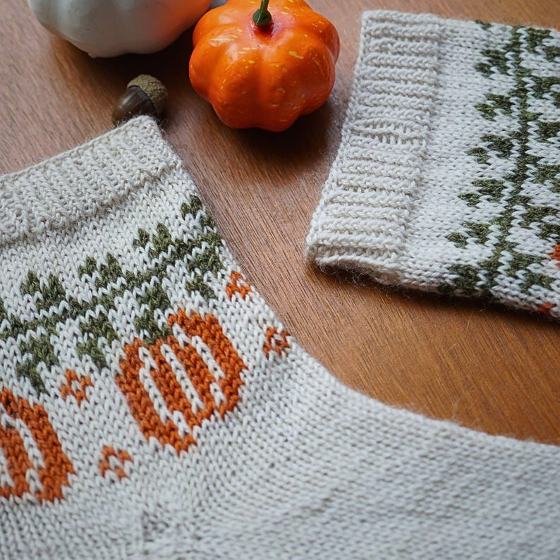 Puede incluir: Primer plano de un calcet&iacute;n y pu&ntilde;o de punto con un dise&ntilde;o de calabaza y hojas en naranja y verde sobre un fondo crema. Tambi&eacute;n se ven calabazas decorativas y una bellota, lo que sugiere un tema oto&ntilde;al.