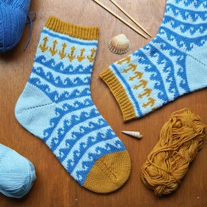 May include: Hand-knitted socks in light blue with a wave pattern and gold accents. The socks feature a nautical design with anchors and a gold cuff and toe. Yarn and knitting needles are also visible.