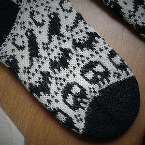 May include: Close-up of a black and white patterned sock. The sock features a repeating geometric design with a skull motif on the toe. The cuff and toe are solid black. The sock is made of a soft, knitted material.
