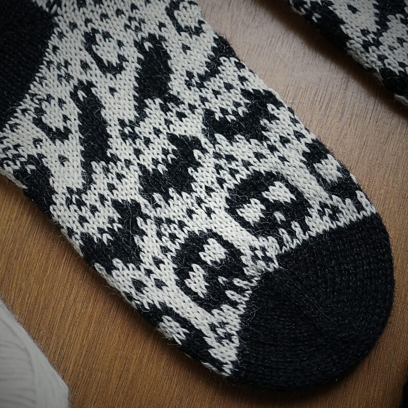 May include: Close-up of a black and white patterned sock. The sock features a repeating geometric design with a skull motif on the toe. The cuff and toe are solid black. The sock is made of a soft, knitted material.