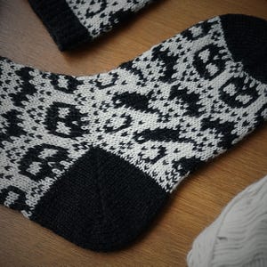 May include: A black and white patterned sock with a matching cuff. The sock features a repeating geometric design in black against a white background. The heel and toe are solid black. A ball of white yarn is visible in the background.