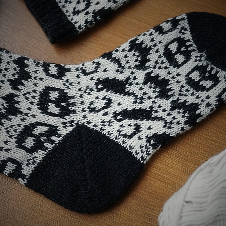 May include: A black and white patterned sock with a matching cuff. The sock features a repeating geometric design in black against a white background. The heel and toe are solid black. A ball of white yarn is visible in the background.