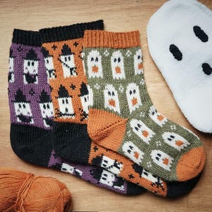 Puede incluir: Tres pares de calcetines de punto con un patrón repetitivo de fantasmas. Un par es morado con puños negros, otro es naranja con puños negros y otro es verde oliva con talón y puntera naranjas. Cada calcetín muestra figuras de fantasmas blancos.