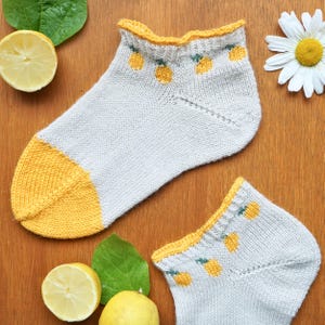 May include: A pair of white ankle socks with yellow accents. The toe and cuff are yellow, and the cuff features a yellow scalloped edge and small yellow lemon designs. The socks are knit and rest on a wooden surface.