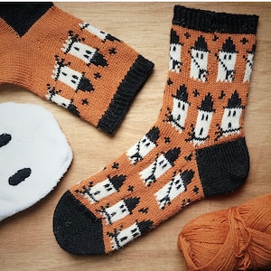 May include: A pair of knitted socks featuring a repeating pattern of white ghosts with black hats on an orange background. The socks have black cuffs, heels, and toes. The design includes small black star accents scattered between the ghost figures.