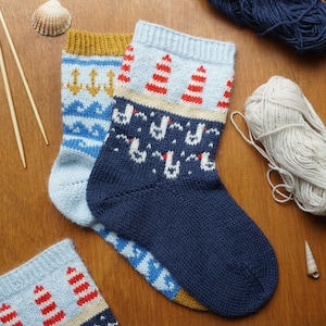 May include: Two pairs of knitted socks with nautical designs. One pair features blue waves and gold anchors on a light blue background, while the other has red and white lighthouses and white seagulls on a navy blue background. The socks are displayed on a wooden surface.