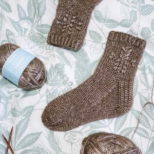 May include: Two brown knitted socks with a snowflake pattern. The socks are lying on a white background with a green floral pattern. Two balls of brown yarn are also in the image.