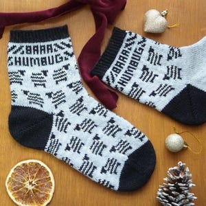 May include: A pair of grey knitted socks with black heels, toes, and cuffs. The socks feature a repeating pattern of black zigzags and the text "BAAA HUMBUG" in black. The socks are displayed with festive decorations on a wooden surface.