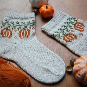 Puede incluir: Un par de calcetines tejidos a mano con un dise&ntilde;o de calabaza. Los calcetines son de color gris claro con patrones de calabaza y vid de color naranja y verde. Una madeja de hilo naranja y peque&ntilde;as calabazas est&aacute;n en el fondo.