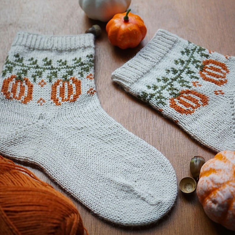 Puede incluir: Un par de calcetines tejidos a mano con un dise&ntilde;o de calabaza. Los calcetines son de color gris claro con patrones de calabaza y vid de color naranja y verde. Una madeja de hilo naranja y peque&ntilde;as calabazas est&aacute;n en el fondo.