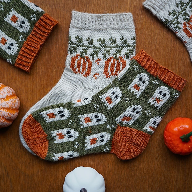 Puede incluir: Calcetines de Halloween tejidos con dise&ntilde;os de fantasmas y calabazas en naranja, blanco y verde. Los calcetines se muestran con peque&ntilde;as calabazas sobre una superficie de madera. Los calcetines tienen pu&ntilde;os y talones naranjas.