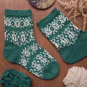 May include: A pair of green knit socks with a white geometric pattern. The socks are displayed on a wooden surface, with a ball of yarn and other decorative elements in the background. The socks appear to be handmade.