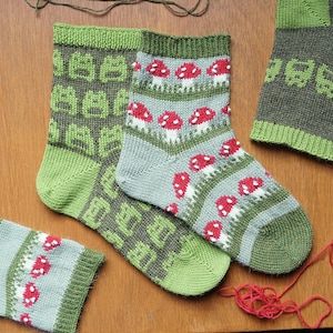 Könnte beinhalten: Zwei Paar gestrickte Socken. Ein Paar hat ein sich wiederholendes Muster aus grünen Quadraten. Das andere hat rote und weiße Pilze auf grauem Hintergrund. Die Socken haben grüne Bündchen, Fersen und Zehen. Aus Wolle gestrickt.