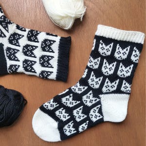 Può includere: Un paio di calzini lavorati a maglia neri e bianchi con un motivo a ripetizione di facce di gatto. I calzini hanno punta e polsino bianchi. Sono visibili anche gomitoli di lana nera e bianca.