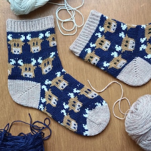 May include: A pair of knitted socks featuring a navy blue design with a repeating pattern of brown and white reindeer. The socks have light brown heels, toes, and cuffs. Yarn is visible in the background.
