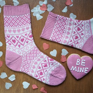 May include: Two pink and white knitted socks with a heart and geometric pattern, alongside a heart-shaped biscuit with "BE MINE" written on it. Heart-shaped confetti is scattered on a wooden surface. The socks have a Valentine's Day theme.