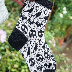 May include: A black and white knitted sock with a skull pattern. The sock has a black cuff, heel, and toe. The main body of the sock features a repeating pattern of white skulls and other shapes on a black background. The sock is held up by a hand.