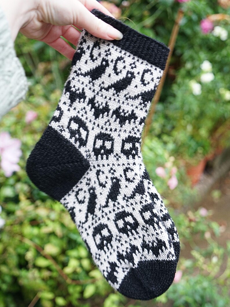 May include: A black and white knitted sock with a skull pattern. The sock has a black cuff, heel, and toe. The main body of the sock features a repeating pattern of white skulls and other shapes on a black background. The sock is held up by a hand.