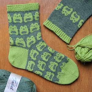 Könnte beinhalten: Handgestrickte Socke in Grün- und Grautönen mit einem geometrischen Muster. Die Socke wird mit einer passenden Manschette und einem Knäuel Wolle präsentiert, um die Handwerkskunst und die verwendeten Materialien zu zeigen. Ferse und Zehen der Socke sind hellgrün.