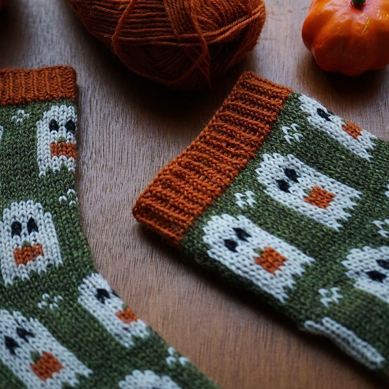 Puede incluir: Un par de calcetines de punto verdes con dise&ntilde;os de fantasmas blancos, pu&ntilde;os naranjas y ojos negros. Una madeja de lana naranja y una peque&ntilde;a calabaza est&aacute;n en el fondo. Los calcetines est&aacute;n sobre una superficie de madera.