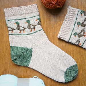 Könnte beinhalten: Eine handgestrickte Socke mit Entenmuster. Die Socke hat einen beigen Körper, eine grüne Ferse und Spitze sowie ein Band mit Enten in Braun, Grün und Orange. Ein passendes Strickmuster ist ebenfalls zu sehen. Die Socke ist aus Garn gefertigt.