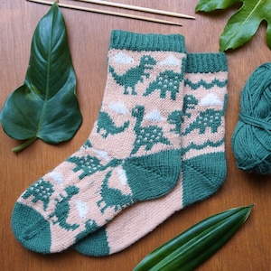 Puede incluir: Un par de calcetines de punto con un diseño de dinosaurios. Los calcetines son principalmente de color rosa claro con diseños de dinosaurios y nubes verdes. Los puños, talones y dedos son verdes. Agujas de tejer y una madeja de lana verde en el fondo.