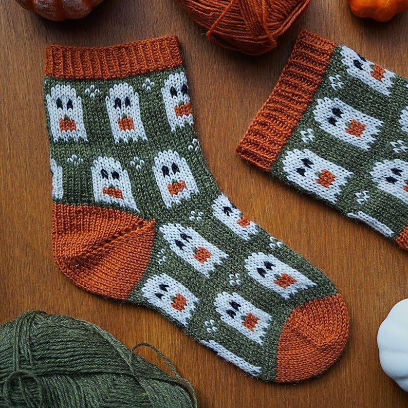 Puede incluir: Un par de calcetines de punto con tem&aacute;tica de Halloween. Los calcetines son de color verde oliva con patrones de fantasmas blancos y detalles en naranja en el pu&ntilde;o, el tal&oacute;n y la puntera. Se muestran sobre una superficie de madera con hilo y calabazas.