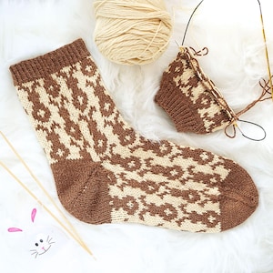 May include: A hand-knitted sock with a brown and cream geometric pattern. The sock is next to a ball of cream-colored yarn and knitting needles. A partially completed sock is also visible, along with a small bunny illustration.