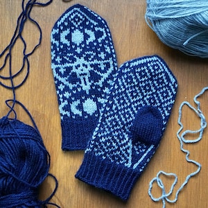 May include: A pair of knitted mittens in navy blue and light blue, featuring intricate geometric patterns. The mittens are displayed on a wooden surface, with balls of yarn in matching colors and loose yarn strands scattered around.
