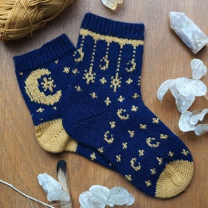 Puede incluir: Un par de calcetines de punto azul marino con detalles dorados. Los calcetines presentan un diseño de luna creciente y estrellas. El talón y la puntera son dorados. Los calcetines se muestran sobre una superficie de madera.