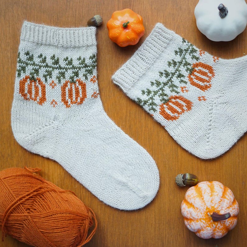 Puede incluir: Un par de calcetines de punto color crema con un dise&ntilde;o de calabaza naranja y vid verde. Los calcetines est&aacute;n decorados con calabazas naranjas y vides verdes. Alrededor, peque&ntilde;as calabazas, bellotas y una madeja de hilo naranja.