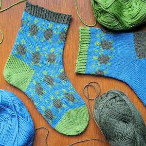 May include: A pair of hand-knitted socks featuring a blue base with a repeating pattern of dark gray turtles with light green accents. The socks have a dark gray ribbed cuff, a light green heel, and a light green toe section, displayed with yarn.