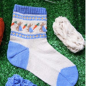 May include: A blue and white knitted sock with a pattern of bunnies and carrots. The sock is on a green surface.