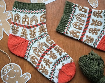 May include: A pair of knitted socks with a festive gingerbread house and tree pattern. The socks feature a white base with brown, red, and green accents. The heel and toe are a solid orange color, and the cuff is green.