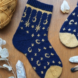 Puede incluir: Calcetines de punto azul marino con un patrón dorado de lunas crecientes y estrellas. El talón y la puntera son dorados. Los calcetines están sobre una superficie de madera con un ovillo de lana y cristales.