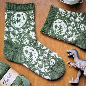 May include: Green and white knitted socks featuring a skull design. The image shows a pair of socks, a matching cuff, and a toy dinosaur on a wooden surface. The yarn label reads "Drops Fabel".