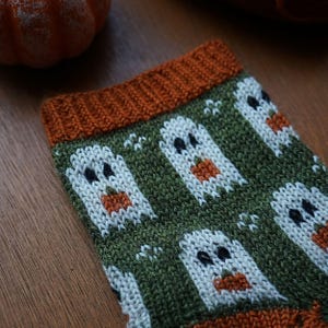 Puede incluir: Primer plano de un calcet&iacute;n de punto con tem&aacute;tica de Halloween. El calcet&iacute;n presenta un fondo verde con dise&ntilde;os de fantasmas blancos y calabazas naranjas. El pu&ntilde;o y la puntera son de color naranja. El calcet&iacute;n est&aacute; hecho de un hilo suave y texturizado.