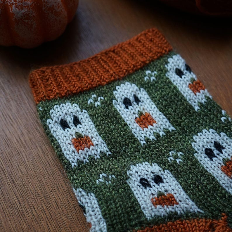 Puede incluir: Primer plano de un calcet&iacute;n de punto con tem&aacute;tica de Halloween. El calcet&iacute;n presenta un fondo verde con dise&ntilde;os de fantasmas blancos y calabazas naranjas. El pu&ntilde;o y la puntera son de color naranja. El calcet&iacute;n est&aacute; hecho de un hilo suave y texturizado.