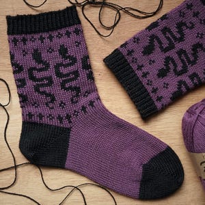 Puede incluir: Un par de calcetines tejidos con un diseño en color morado y negro. Los calcetines presentan un patrón repetitivo de serpientes negras y pequeñas formas de estrella sobre un fondo morado, con puños, talones y punteras en negro sólido.