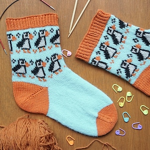 May include: A pair of hand-knitted socks with a penguin pattern. The socks feature a light blue body, orange heel and toe, and orange cuff. The penguin design is black and white. Knitting needles, yarn, and stitch markers are also visible.