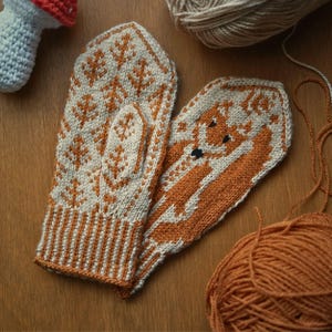 May include: A pair of knitted mittens with a fox design. One mitten is brown with a white fox, and the other is white with a brown fox. The mittens are made with a fair isle pattern.