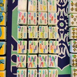 May include: A set of white Mahjong tiles arranged in rows, featuring colorful dotted numerical designs from three to nine, alongside tiles with gold fleur-de-lis symbols and tiles labeled Joker, all displayed on a blue and green patterned mat.