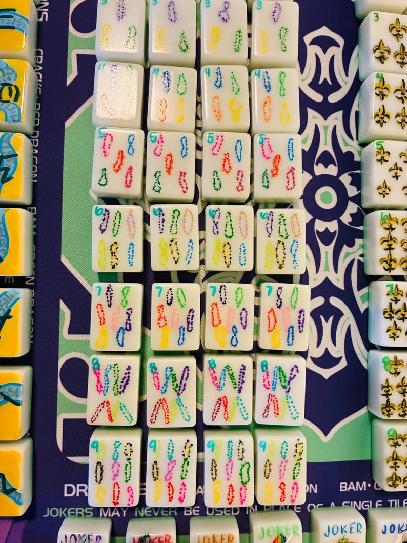 May include: A set of white Mahjong tiles arranged in rows, featuring colorful dotted numerical designs from three to nine, alongside tiles with gold fleur-de-lis symbols and tiles labeled Joker, all displayed on a blue and green patterned mat.