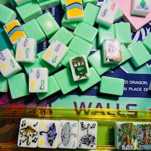 May include: A collection of white mahjong tiles with various hand-painted designs including umbrellas, trees, and text, resting on a game board. The tiles feature green backs with a geometric pattern and are arranged in a scattered pile and a rack.