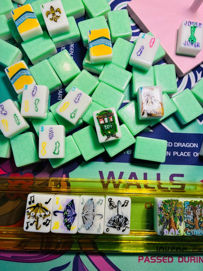 May include: A collection of white mahjong tiles with various hand-painted designs including umbrellas, trees, and text, resting on a game board. The tiles feature green backs with a geometric pattern and are arranged in a scattered pile and a rack.